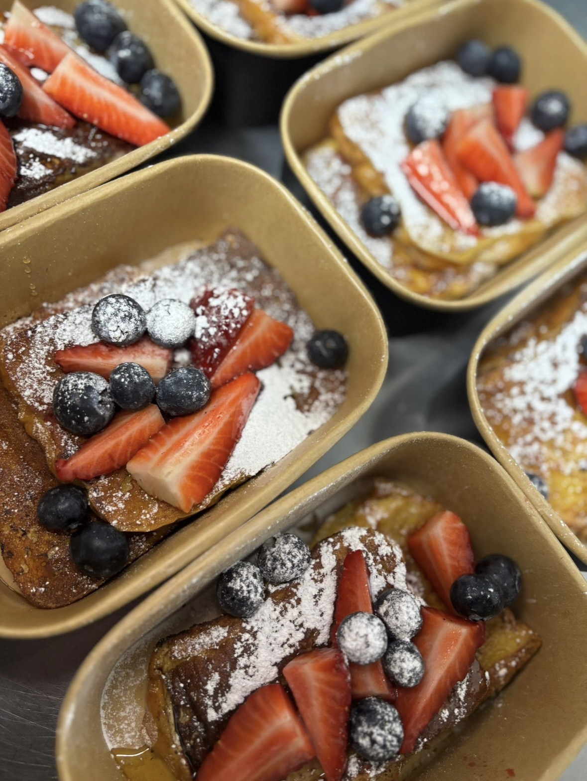 French toast topped with strawberries, blueberries and icing sugar in takeaway trays